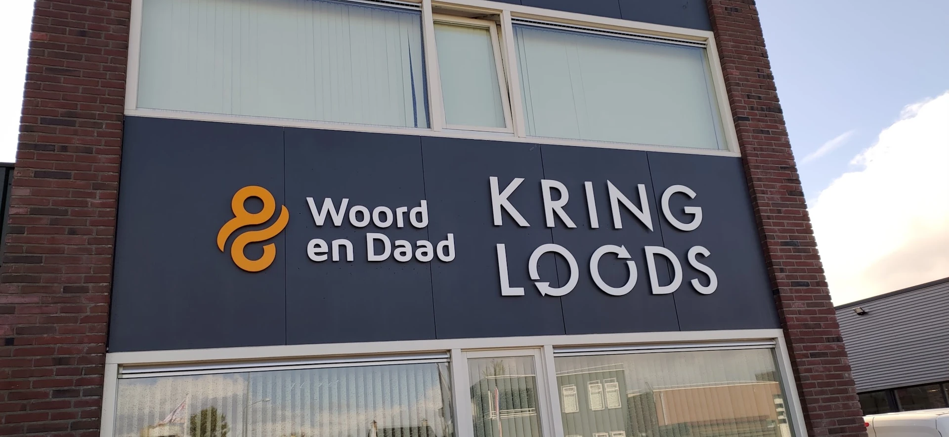 Gevel van kringloopwinkel met donkerblauwe uithangtekens met logo en naam Woord en Daad / Kringloods