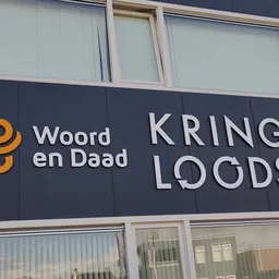 Gevel van kringloopwinkel met donkerblauwe uithangtekens met logo en naam Woord en Daad / Kringloods