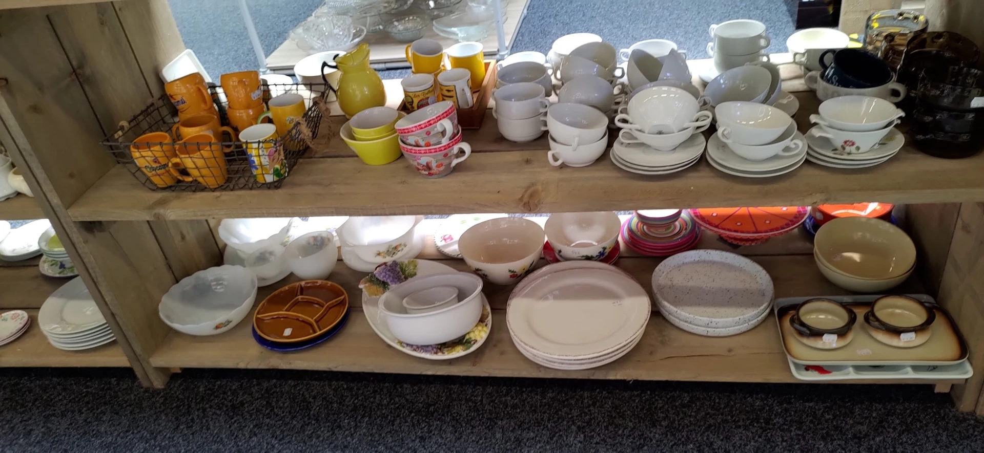 Serviesgoed en borden uitgestald op twee houten schappen, waaronder witte en gekleurde kopjes, schalen en borden
