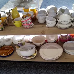 Serviesgoed en borden uitgestald op twee houten schappen, waaronder witte en gekleurde kopjes, schalen en borden