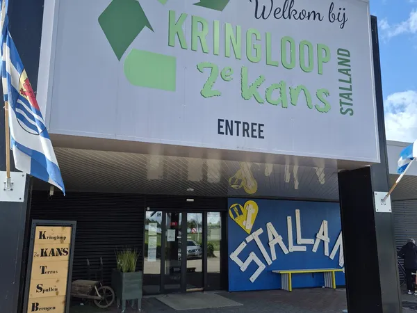Voorgevel van kringloopwinkel met groot welkomstbord, groene logoletters, glazen entree en blauwe decoratief element