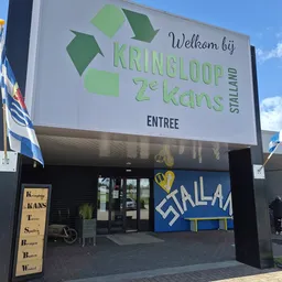 Voorgevel van kringloopwinkel met groot welkomstbord, groene logoletters, glazen entree en blauwe decoratief element