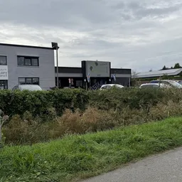 Voorkant van kringloopwinkel aan Ceciliaweg met witte woning links en zwart gebouw met groene accenten, omgeven door begroeii