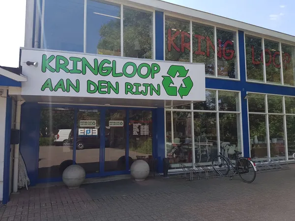 Gevel van kringloopwinkel met groen logo 'KRINGLOOP AAN DEN RIJN' met recyclesymbool, blauwe pui, grote ramen en ingang