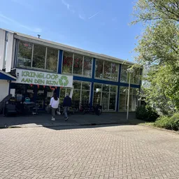 Voorkant van een moderne kringloopwinkel met grote ramen en blauwe luifel, enkele bezoekers zichtbaar.