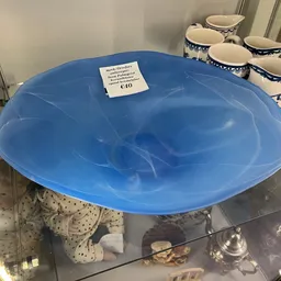 Blauwe ovale serveerschaal met prijskaartje onder vitrineglas, naast blauwe en witte serviesgoed