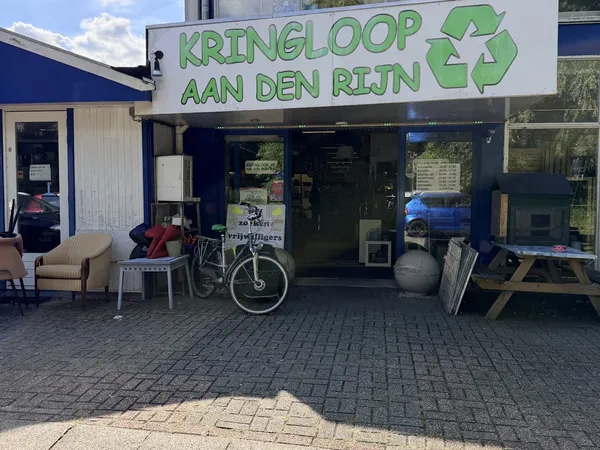 Winkelgevel van Kringloop aan den Rijn met groene letters, etalageruiten en ingang, fietsen en meubels voor de deur