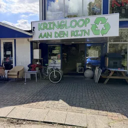 Winkelgevel van Kringloop aan den Rijn met groene letters, etalageruiten en ingang, fietsen en meubels voor de deur