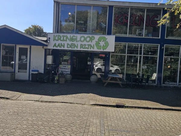 Gevel van kringloopwinkel Kringloop aan den Rijn met groene letters en logo, winkelramen met uitstallingen, ingang met OPEN-b