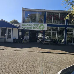 Gevel van kringloopwinkel Kringloop aan den Rijn met groene letters en logo, winkelramen met uitstallingen, ingang met OPEN-b