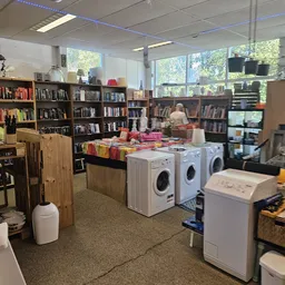 Winkelinterieur met boeken in kasten, wasmachines en was aan een rek, grote ramen met uitzicht op groen