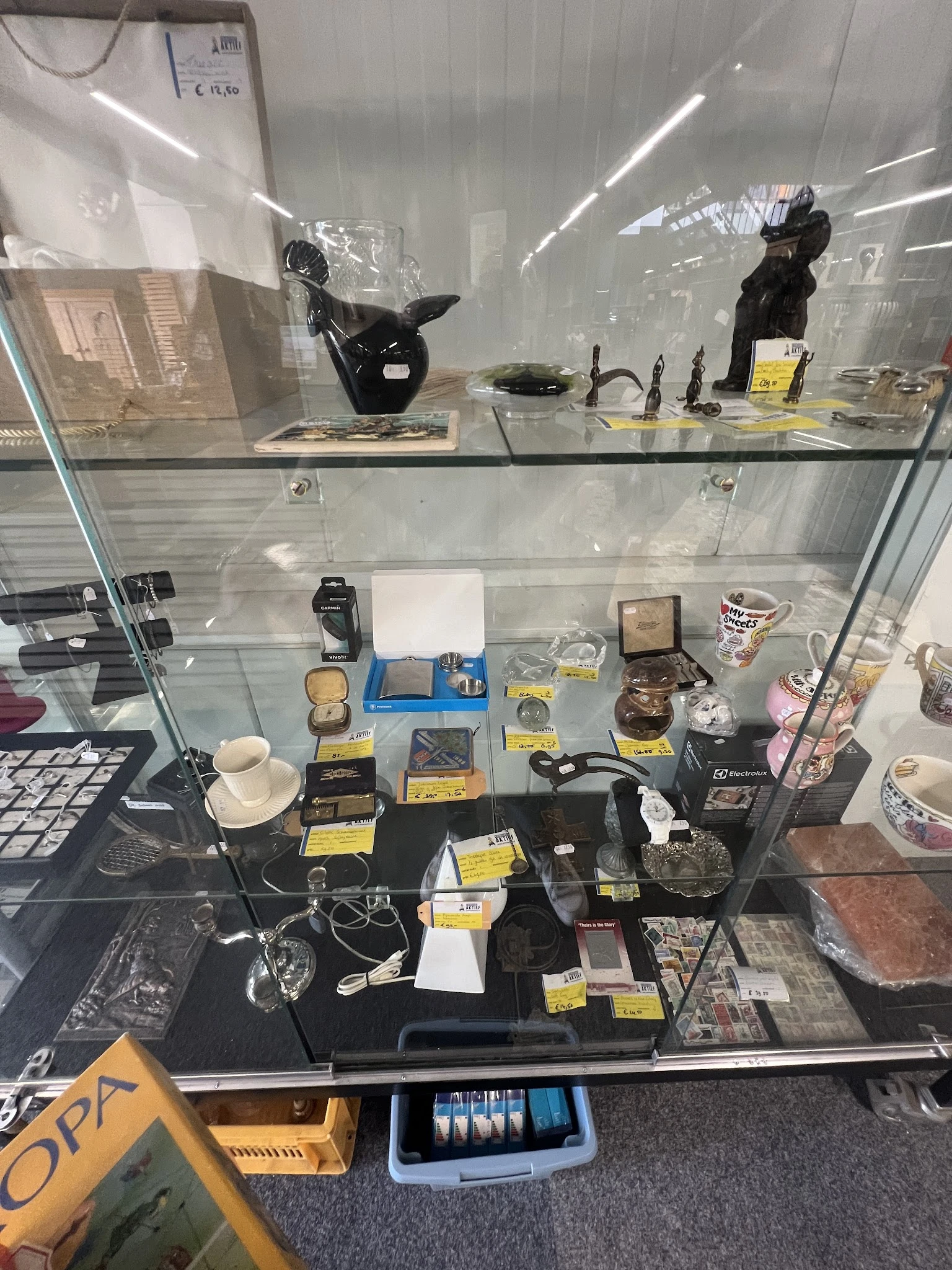 Glazen vitrinekast met diverse tweedehands artikelen zoals servies, accessoires en decoratieve items met gele prijskaartjes