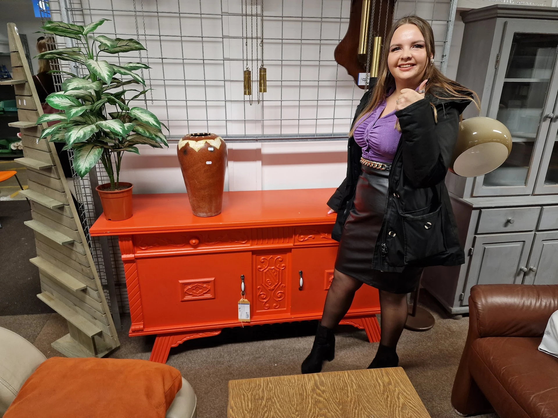 Vrouw in winterjas poseert naast een felrood dressoir in een kringloopwinkel