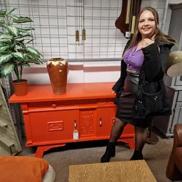 Vrouw in winterjas poseert naast een felrood dressoir in een kringloopwinkel