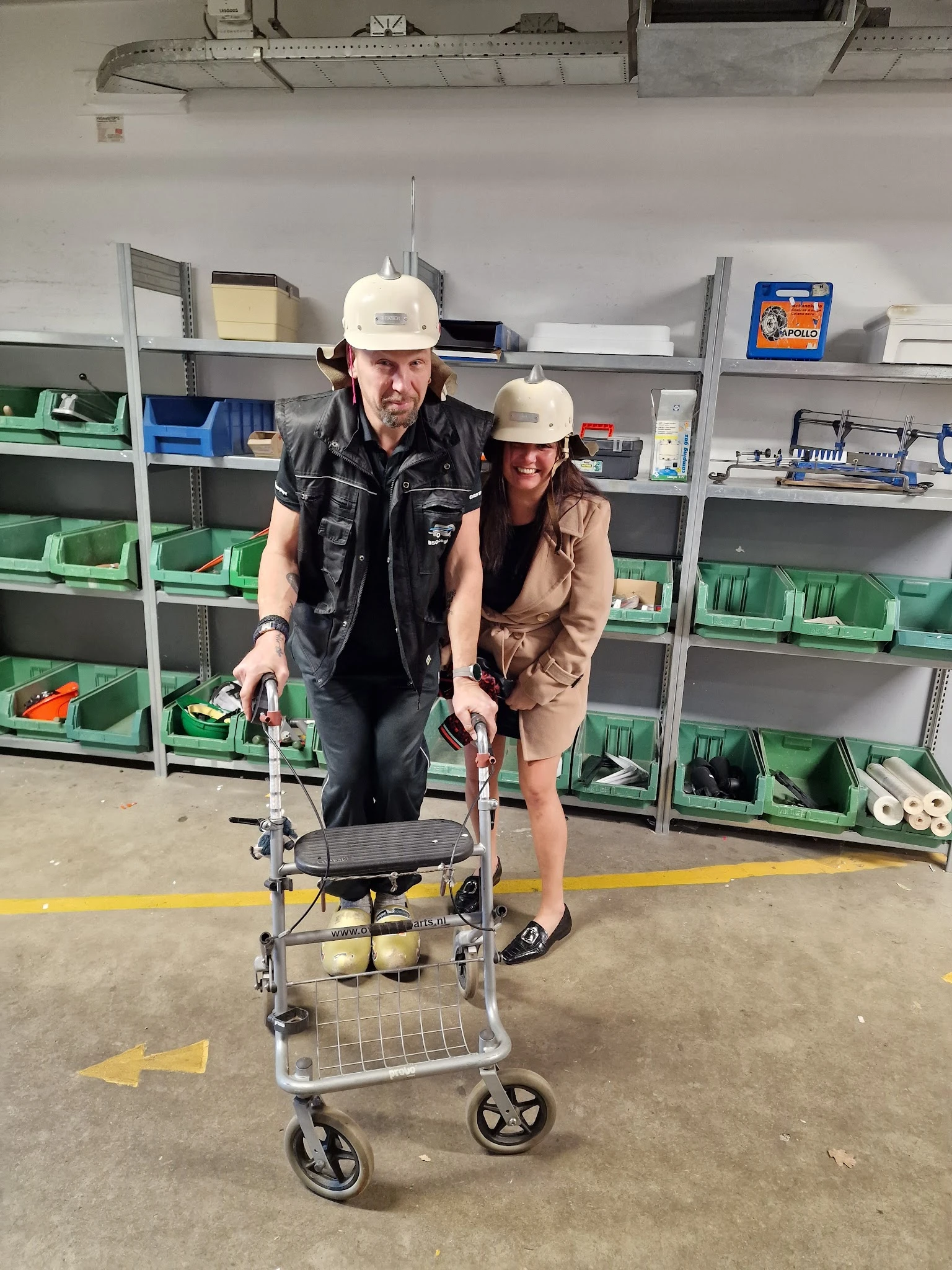 Twee medewerkers in werkkleding met witte helmen poseren met een looprekje in een magazijn met groene stellingen op de achter