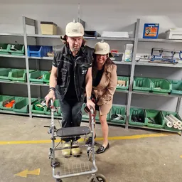 Twee medewerkers in werkkleding met witte helmen poseren met een looprekje in een magazijn met groene stellingen op de achter
