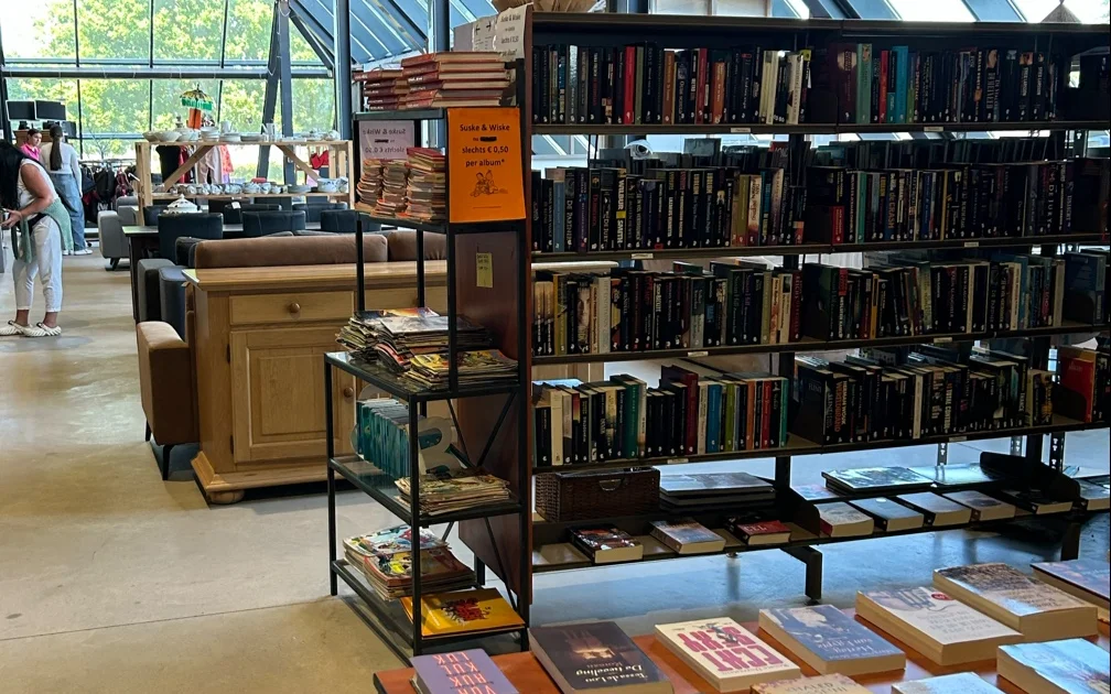 Winkelinterieur met boekenrekken, houten meubels en glazen dak; klanten in de achtergrond