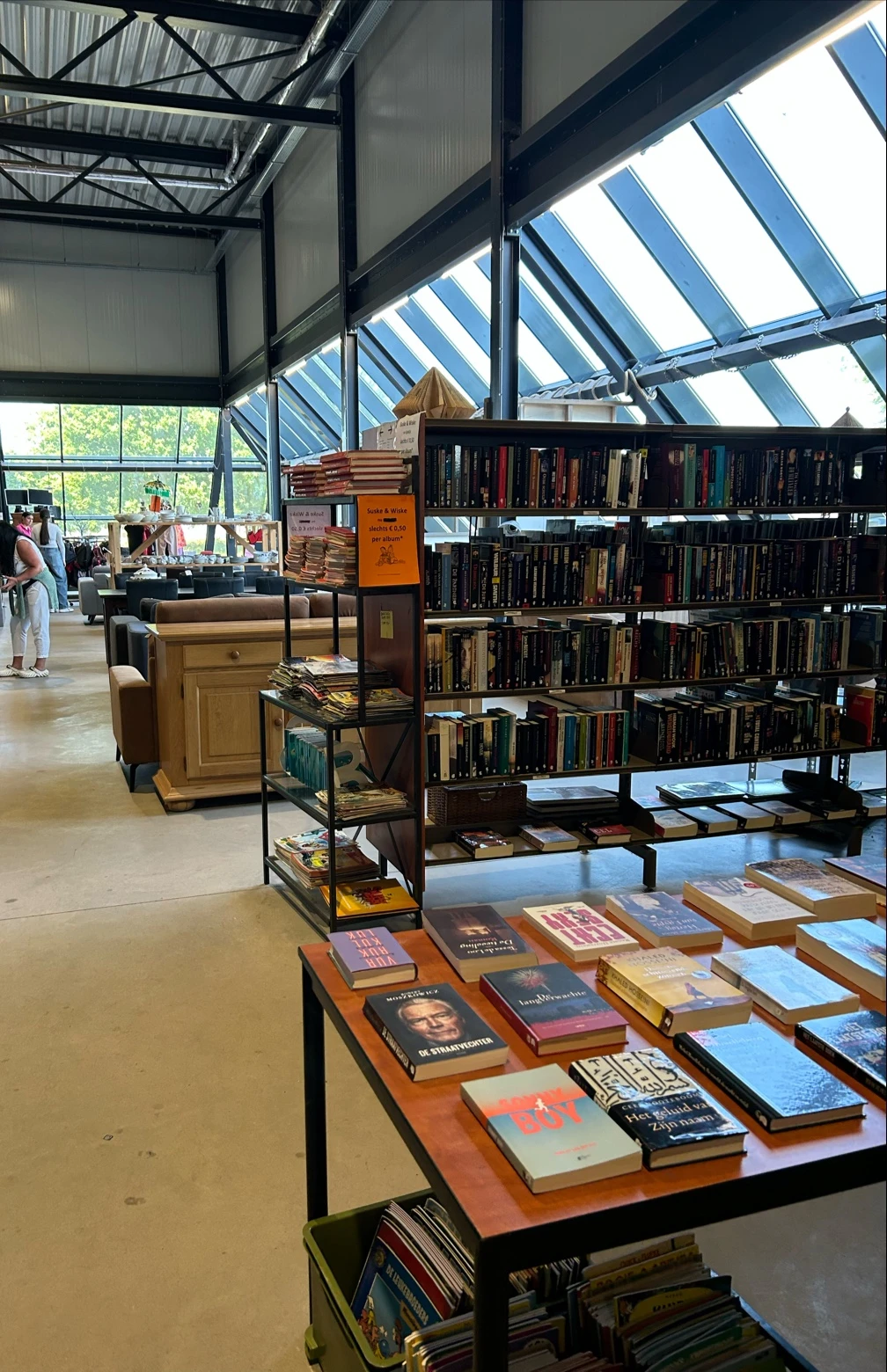 Winkelinterieur met boekenrekken, houten meubels en glazen dak; klanten in de achtergrond