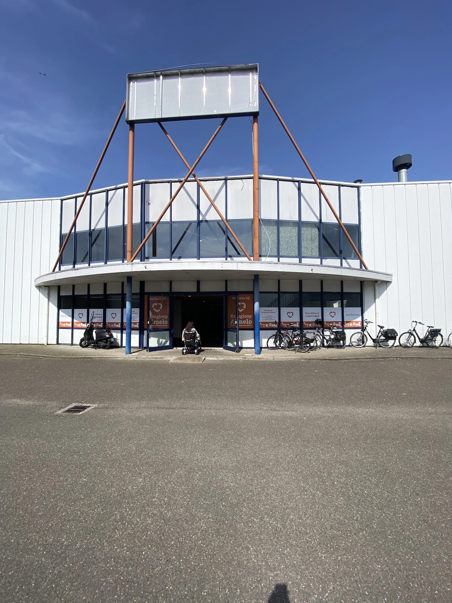 Voorkant van het XL kringloopwarenhuis met karakteristieke ronde vorm, grote ramen en stalen constructie, fietsen geparkeerd 