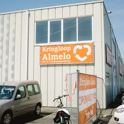 Gevel van kringloopwinkel met oranje bord met hartlogo en tekst 'Kringloop Almelo het XL kringloopwarenhuis'