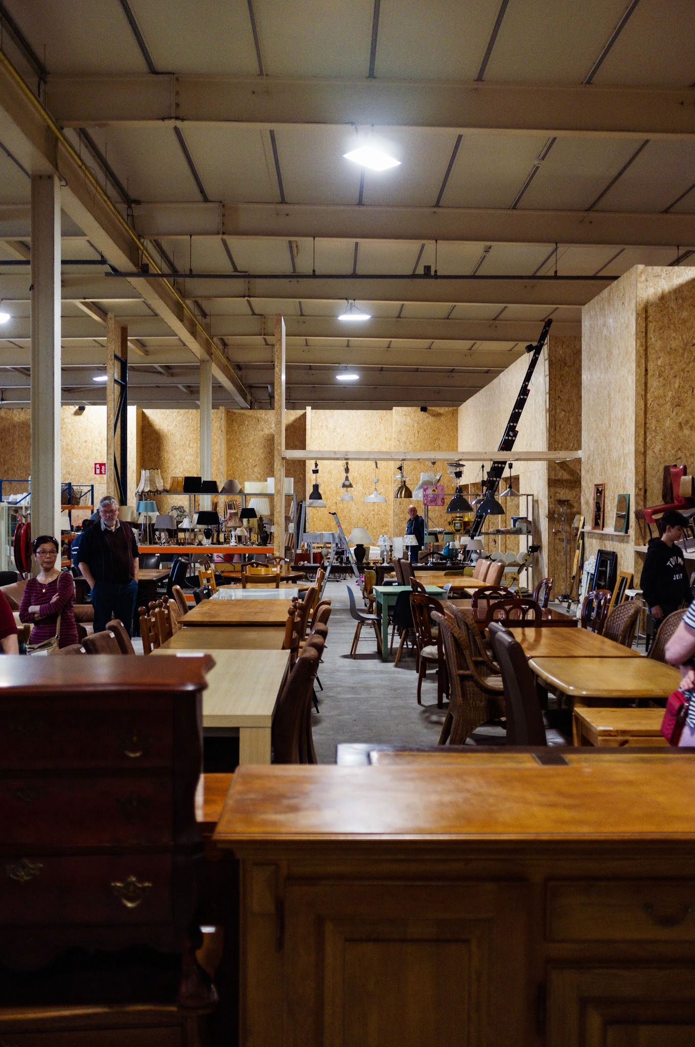 Ruim winkelbinnenruimte met houten tafels, stoelen, en diverse tweedehands spullen op stellingen en aan het plafond