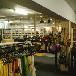 Winkelvloer met kledingrekken, tassen en diverse spullen op tafels, stellingen met boeken op de achtergrond