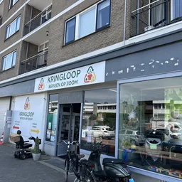 Gevel van kringloopwinkel met blauw uithangbord, grote ramen en ingang aan straatkant