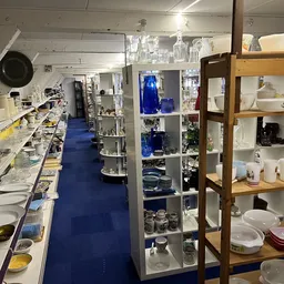 Winkelinterieur met blauwe vloer, kasten met servies en huishoudelijke spullen, goederen op tafels langs de zijkanten