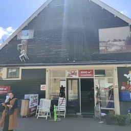 Gevel van kringloopwinkel met donkergroene houten wanden, witte ramen en ingang met spullen buiten uitgestald