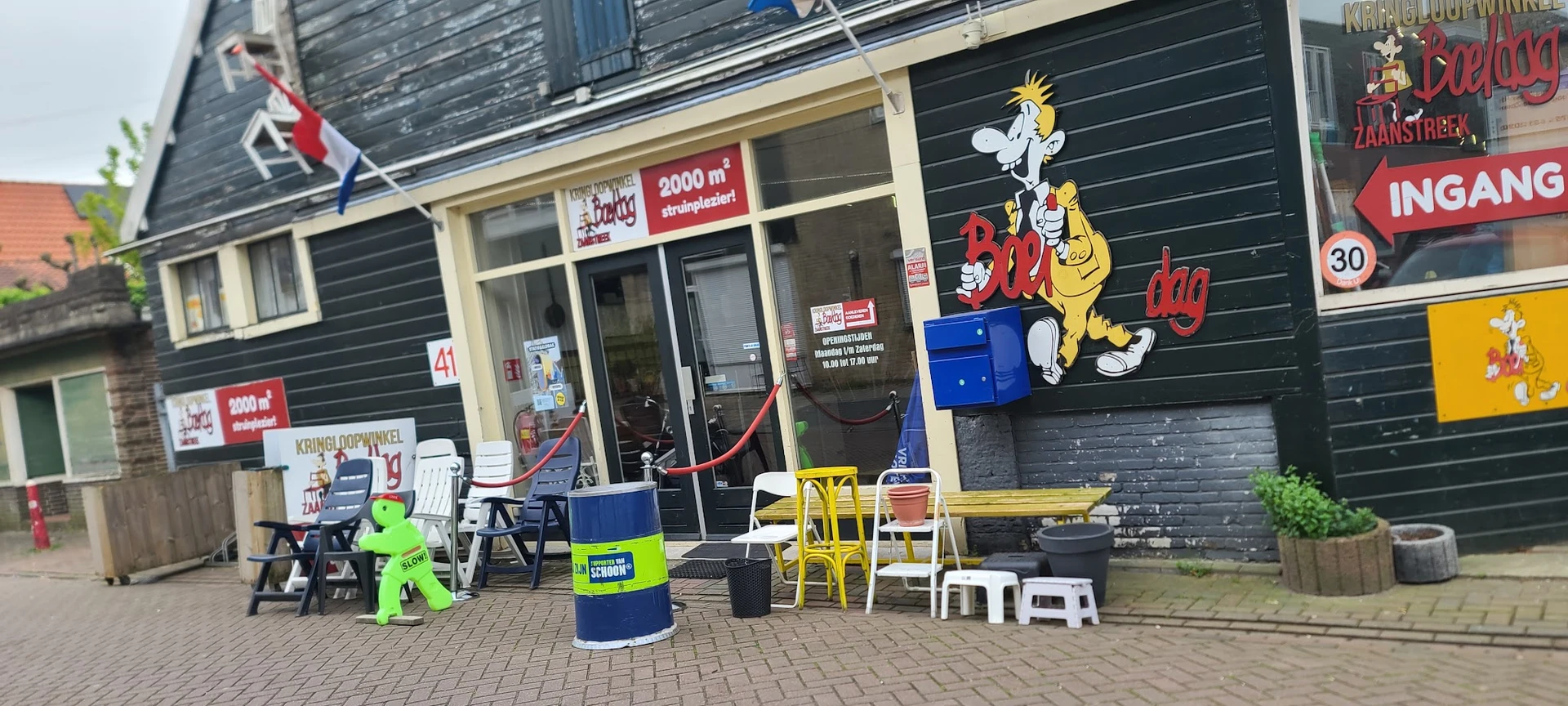 Voorkant kringloopwinkel met etalage, rode deur, speelgoed en meubels buiten, gele stoelen en decoratie