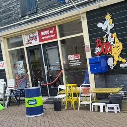 Voorkant kringloopwinkel met etalage, rode deur, speelgoed en meubels buiten, gele stoelen en decoratie