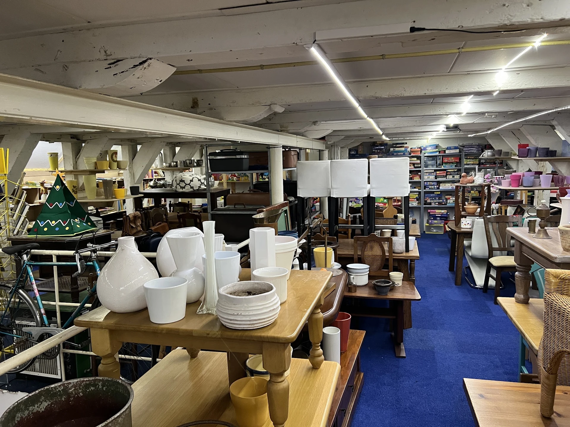 Winkelvloer van kringloopwinkel met blauwe loper, diverse huishoudspullen, servies en meubelstukken uitgestald