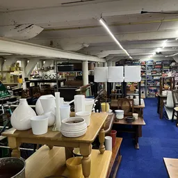 Winkelvloer van kringloopwinkel met blauwe loper, diverse huishoudspullen, servies en meubelstukken uitgestald