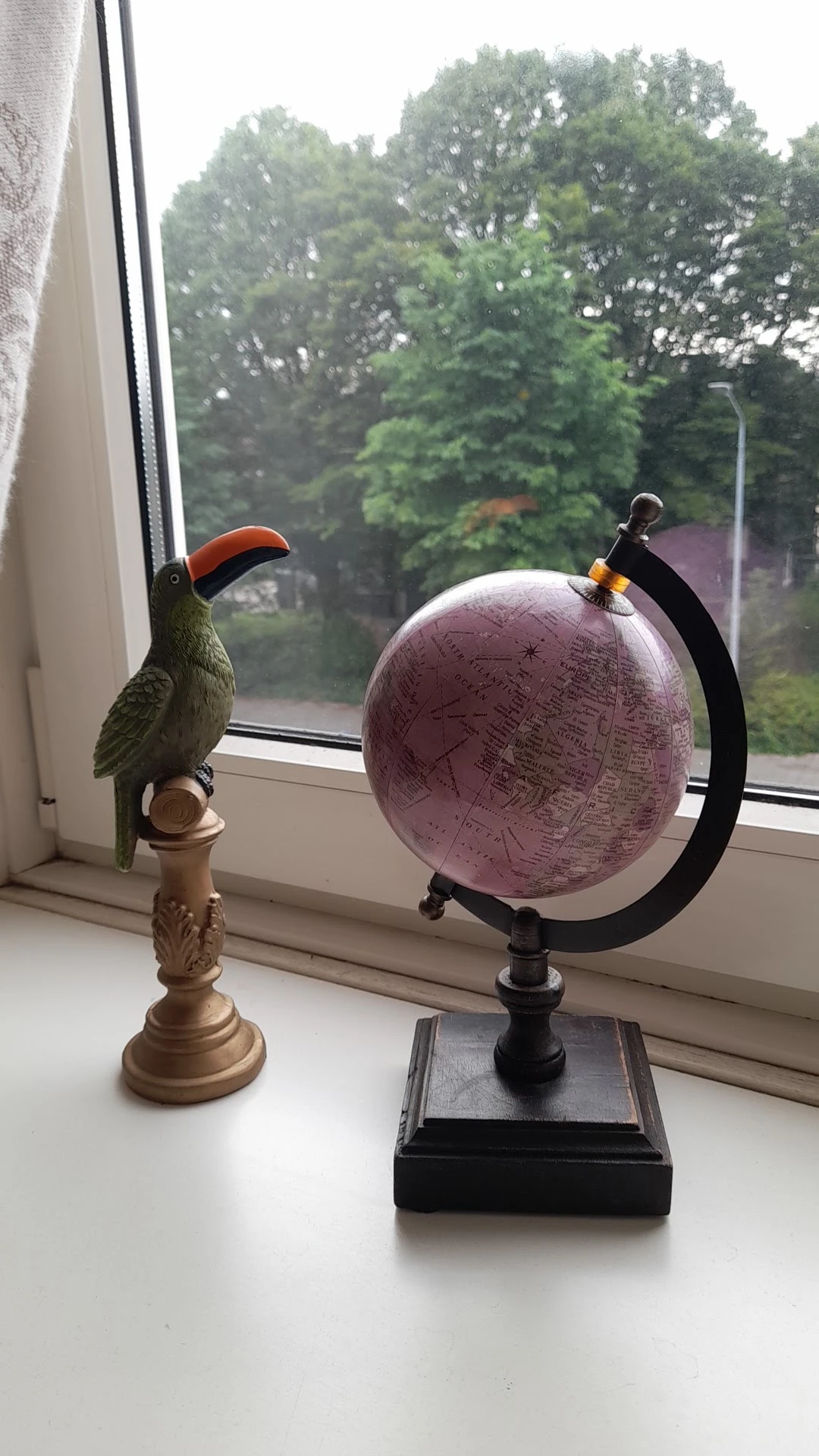 Decoratief vogelfiguur met rood en groen gevederte naast een paarse wereldbol op een vensterbank