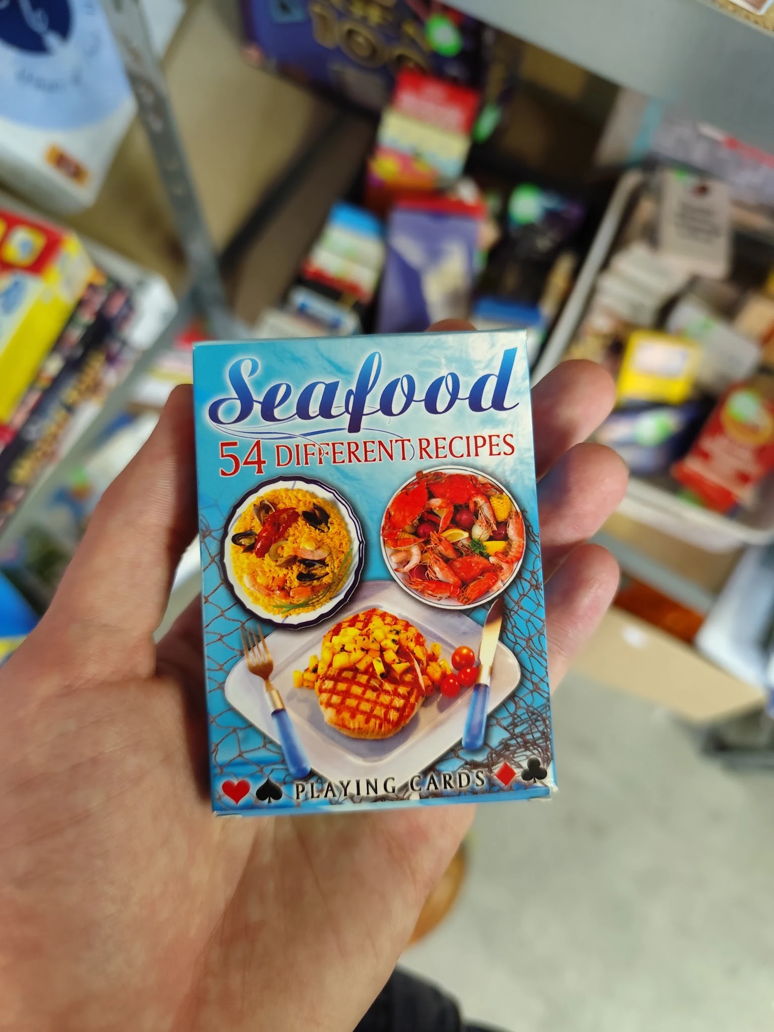 Speelkaartenset met zeevrouchtrecepten in een hand, op de achtergrond onscherpe winkelrek.