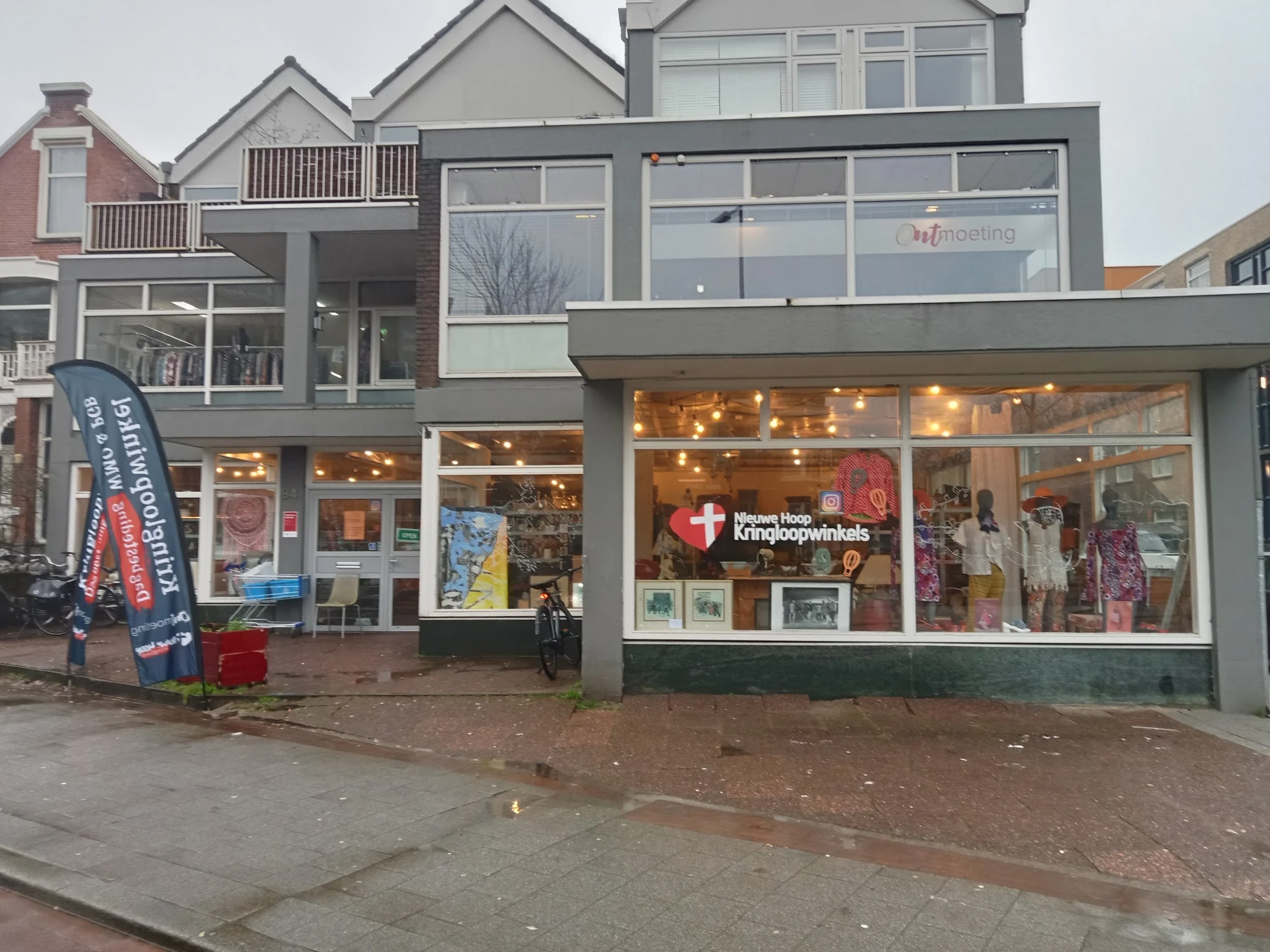 Gevel van kringloopwinkel met grote ramen, verlicht interieur met uitgestalde spullen, winkelnaam zichtbaar, rode deur links