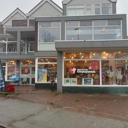 Gevel van kringloopwinkel met grote ramen, verlicht interieur met uitgestalde spullen, winkelnaam zichtbaar, rode deur links
