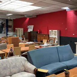 Winkelvloer met diverse tweedehands meubels: blauwe bank, grijze fauteuil, houten tafels en witte kasten tegen rode wand