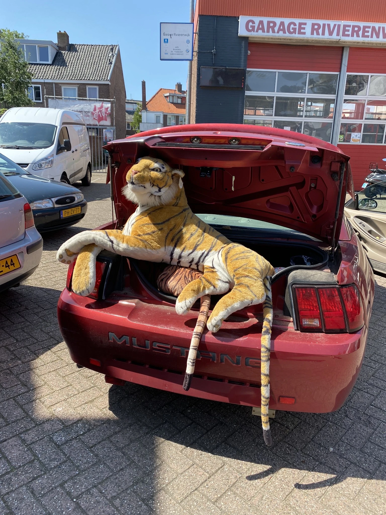 Een grote gele tijger-pop zit in de open kofferbak van een rode auto op een parkeerterrein