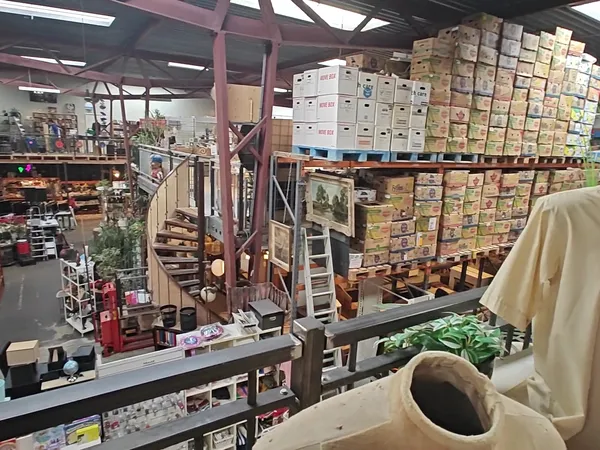 Overzicht van kringloopwinkel met stellingen vol dozen, dozen gestapeld op hoogte, trap en roze balken, diverse spullen op we