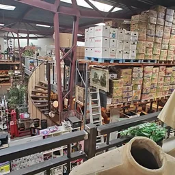 Overzicht van kringloopwinkel met stellingen vol dozen, dozen gestapeld op hoogte, trap en roze balken, diverse spullen op we