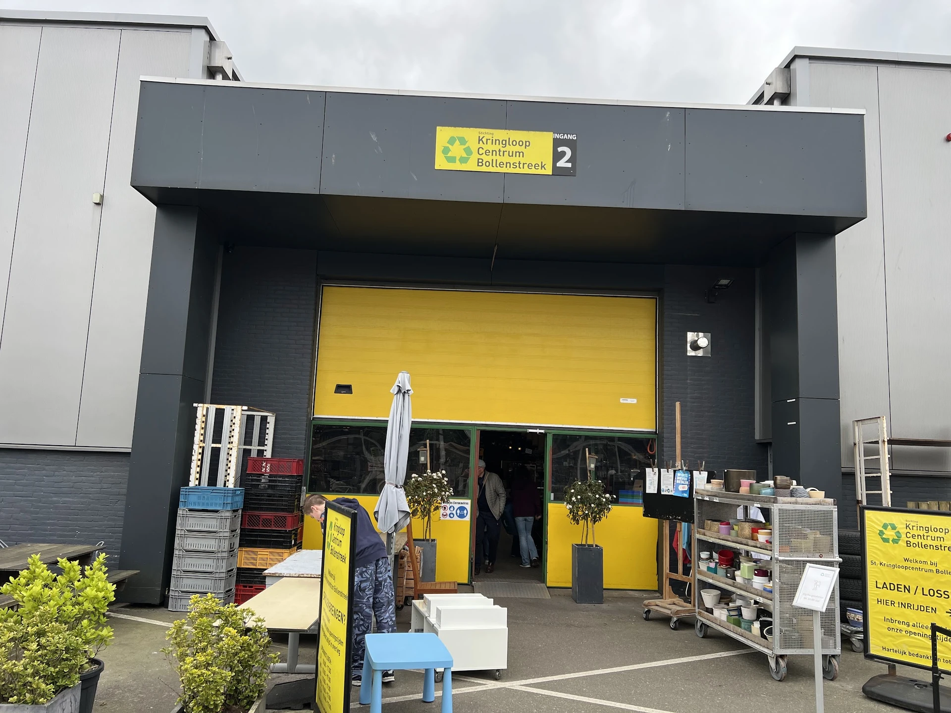 Voorkant van kringloopwinkel met geel en grijs gevelkleur, grote gele roldeur, nummer 2 zichtbaar, enkele spullen buiten uitg