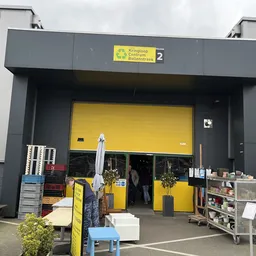 Voorkant van kringloopwinkel met geel en grijs gevelkleur, grote gele roldeur, nummer 2 zichtbaar, enkele spullen buiten uitg