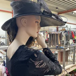 Etalagepop gekleed in zwart kanten kleding en zwarte cowboyhoed, op achtergrond rekken met kleding