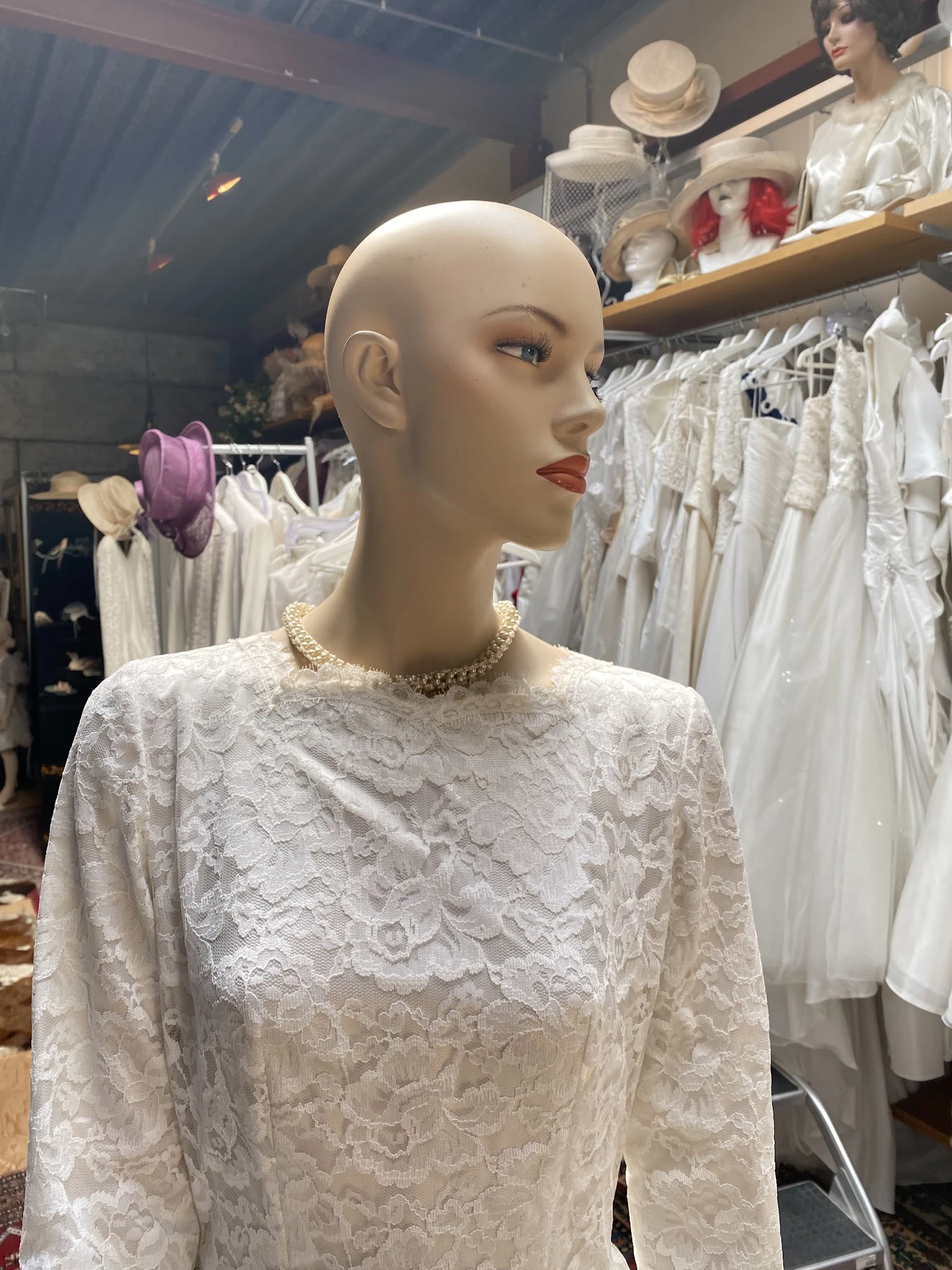 Etalagepop in witte kanten jurk met parelketting, omgeven door rekken met trouwjurken