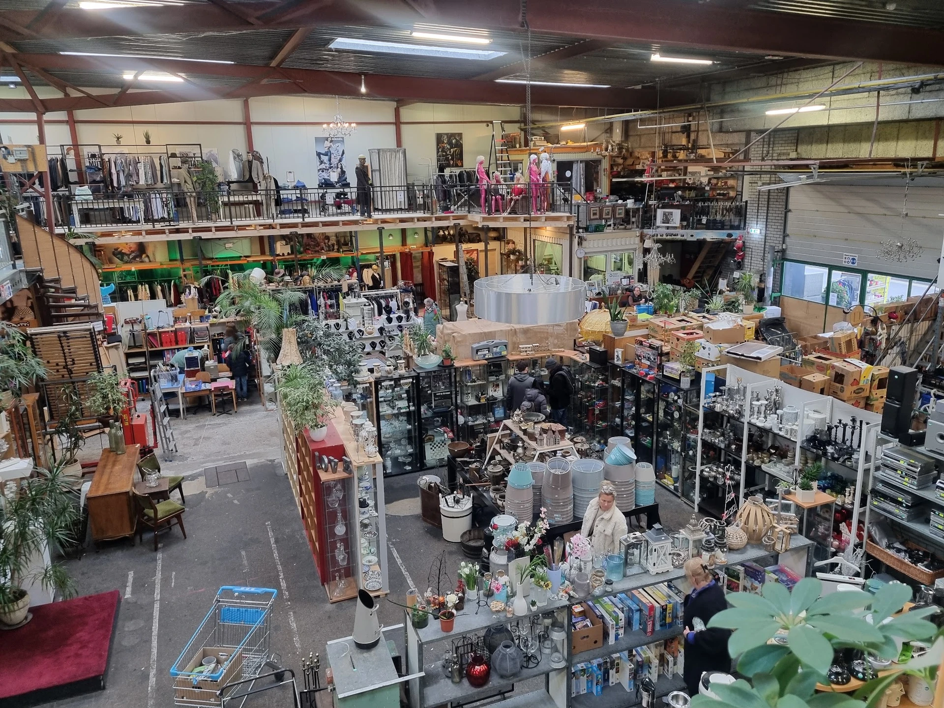 Overzicht van kringloopwinkel met diverse afdelingen, stellingen met spullen, planten en meubilair op meerdere niveaus