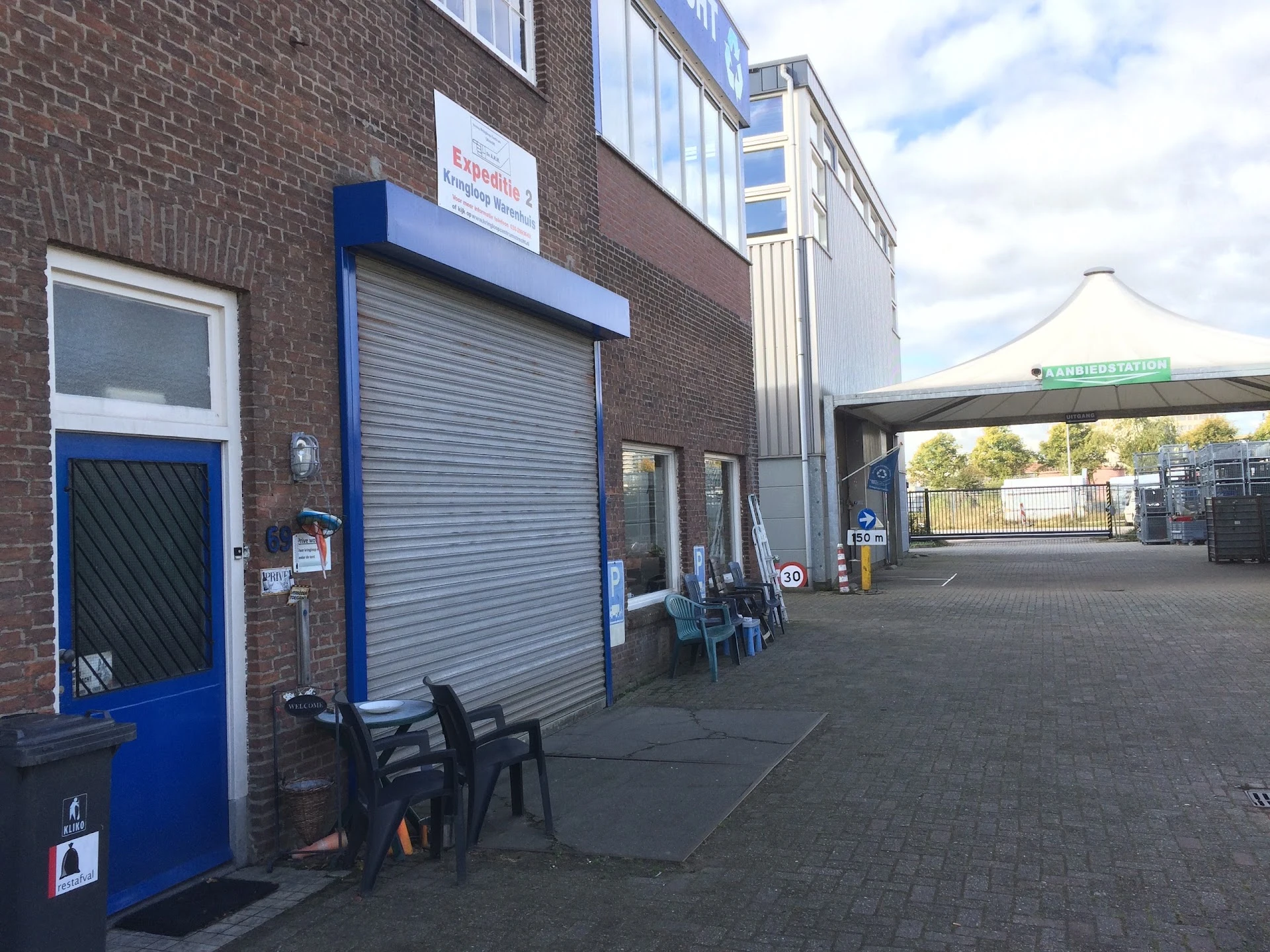 Bakstenen gevel van kringloopwinkel met blauwe deur, rolluik en stellingen buiten