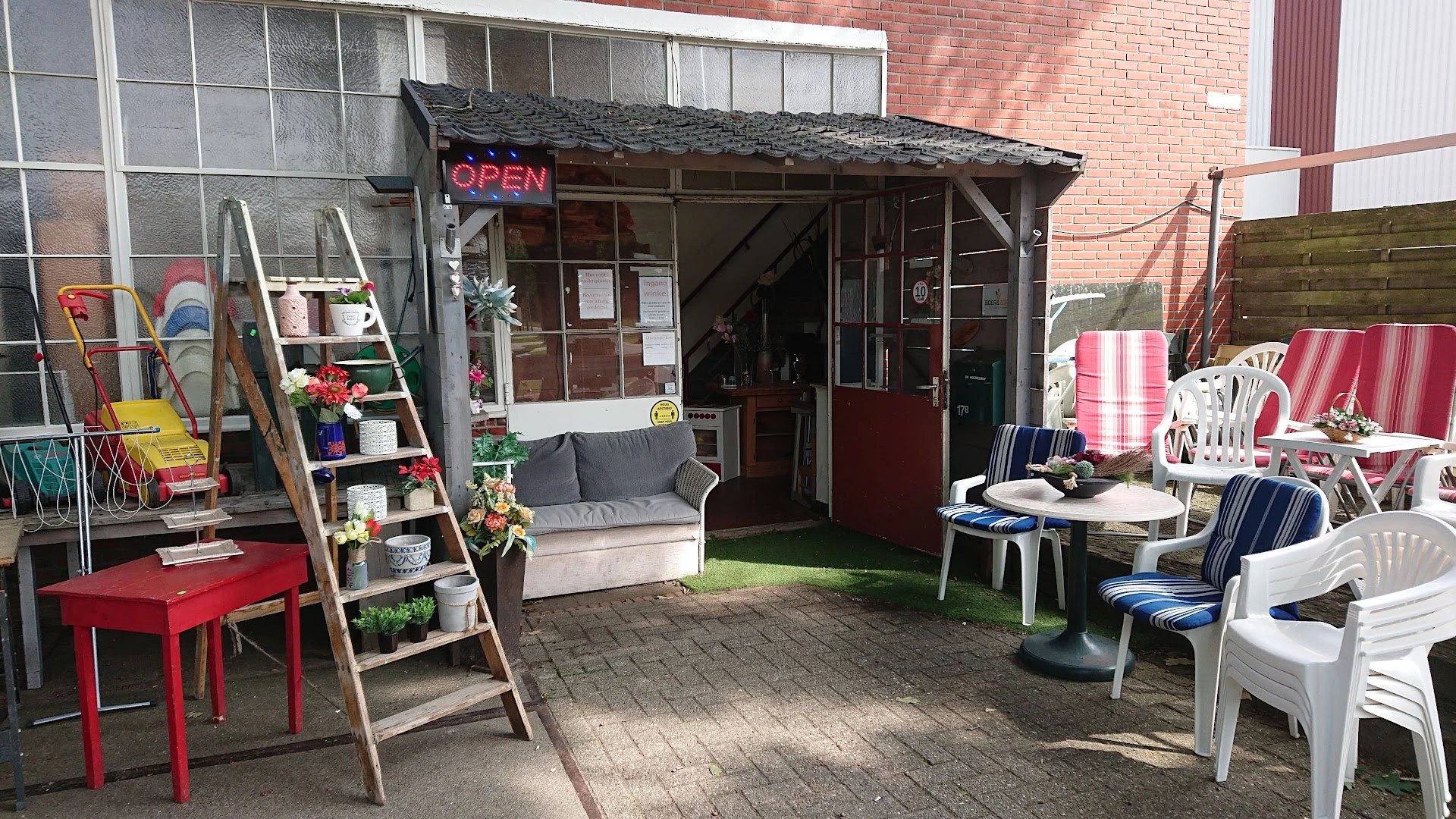 Voorkant van kringloopwinkel met rode deur, OPEN-bord, en tuinmeubelen op terras