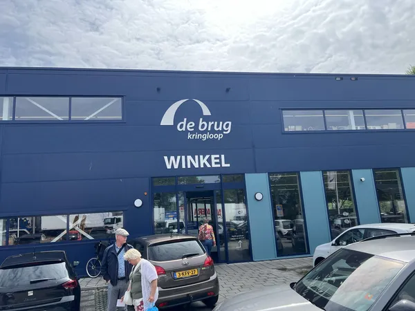 Blauwe gevel van kringloopwinkel De Brug met ingang en parkeerplaats ervoor