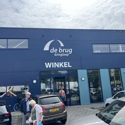 Blauwe gevel van kringloopwinkel De Brug met ingang en parkeerplaats ervoor